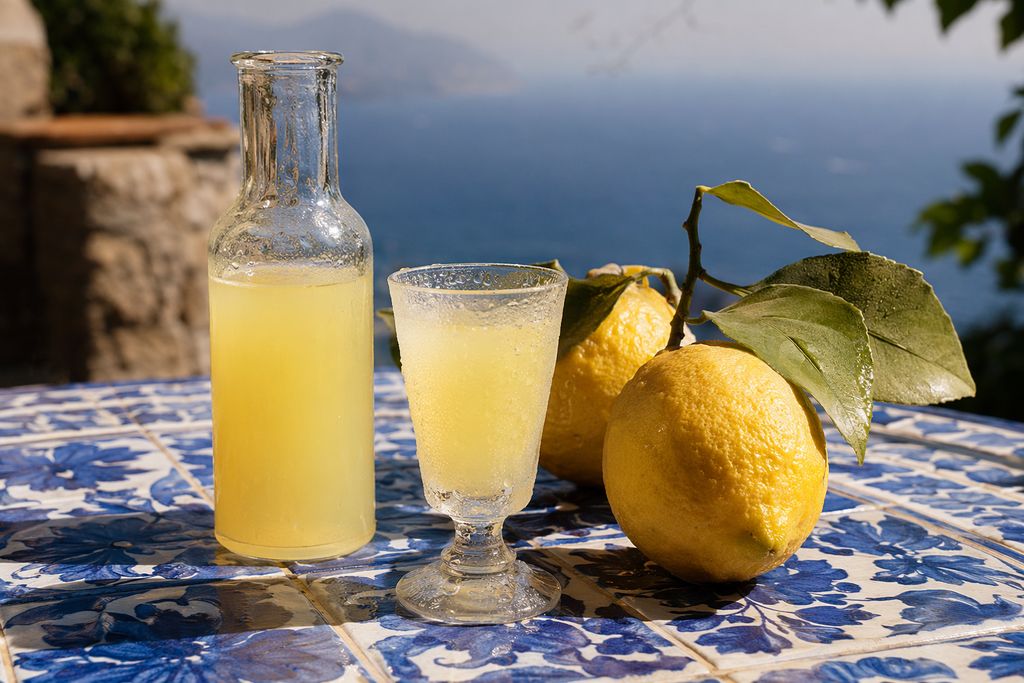 Een beslagen karaf en een ijskoud glas limoncello op een tafel van blauwe tegels, met citroenen en de zee op de achtergrond
