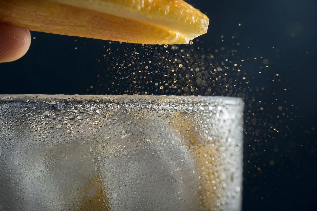 Macro-opname van citroenschil die boven een beslagen, ijskoud glas wordt uitgeknepen, met druppeltjes citrusolie in het zonlicht